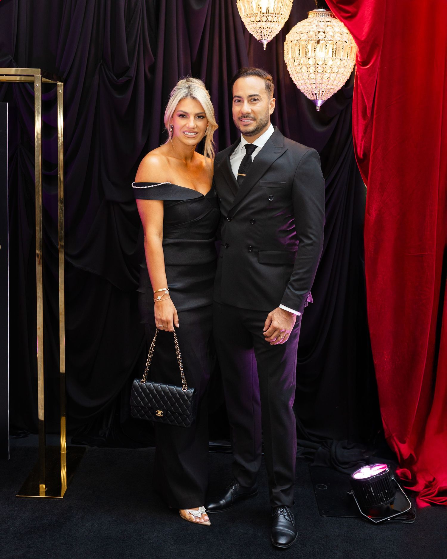 A couple in formal black attire poses elegantly against a backdrop with red curtains and a crystal chandelier.