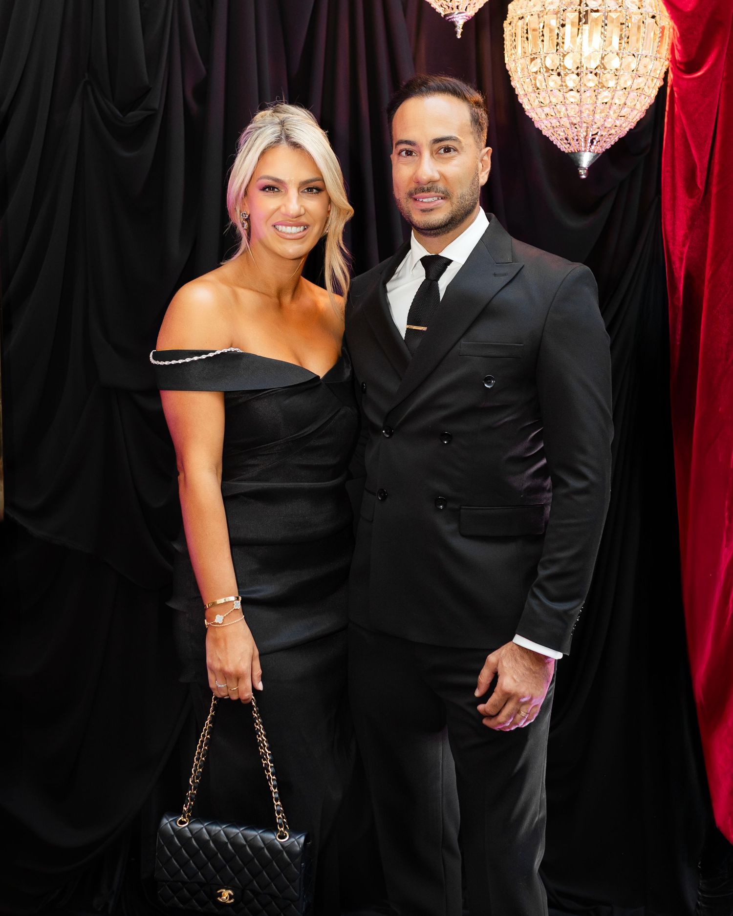 A couple in formal attire poses at an elegant event with chandeliers and dark drapes in the background.
