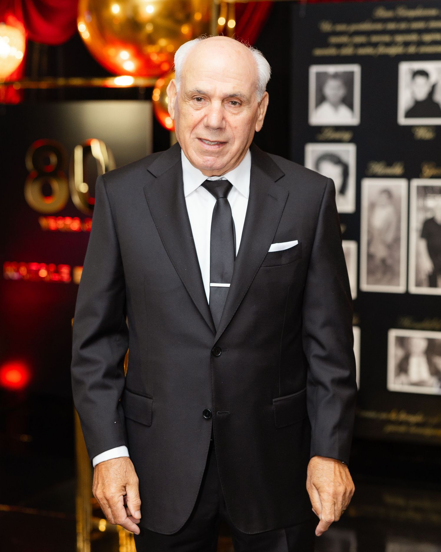 A senior man wearing a black suit and tie stands in front of a dimly lit wall with framed photographs.