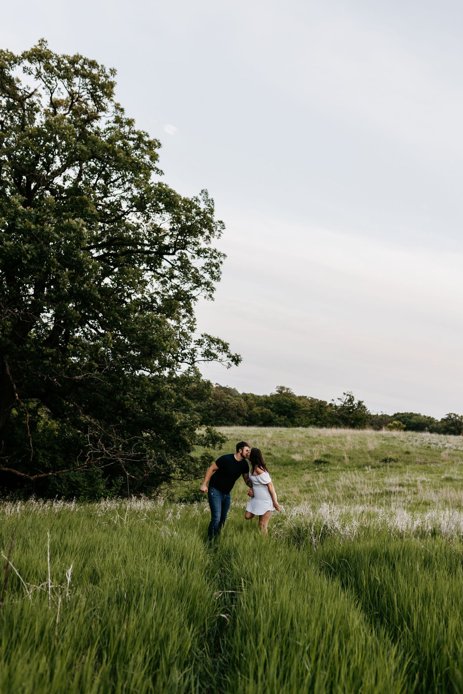 Two figures walk hand-in-hand through a lush meadow surrounded by tall green grass and trees on a cloudy day.