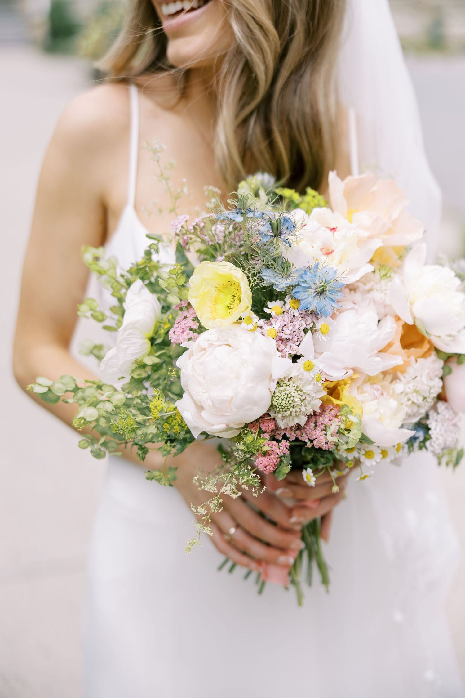 A delicate pastel bridal bouquet with peonies, roses and wildflowers against a white dress at Silver Bay YMCA in Lake George, NY.