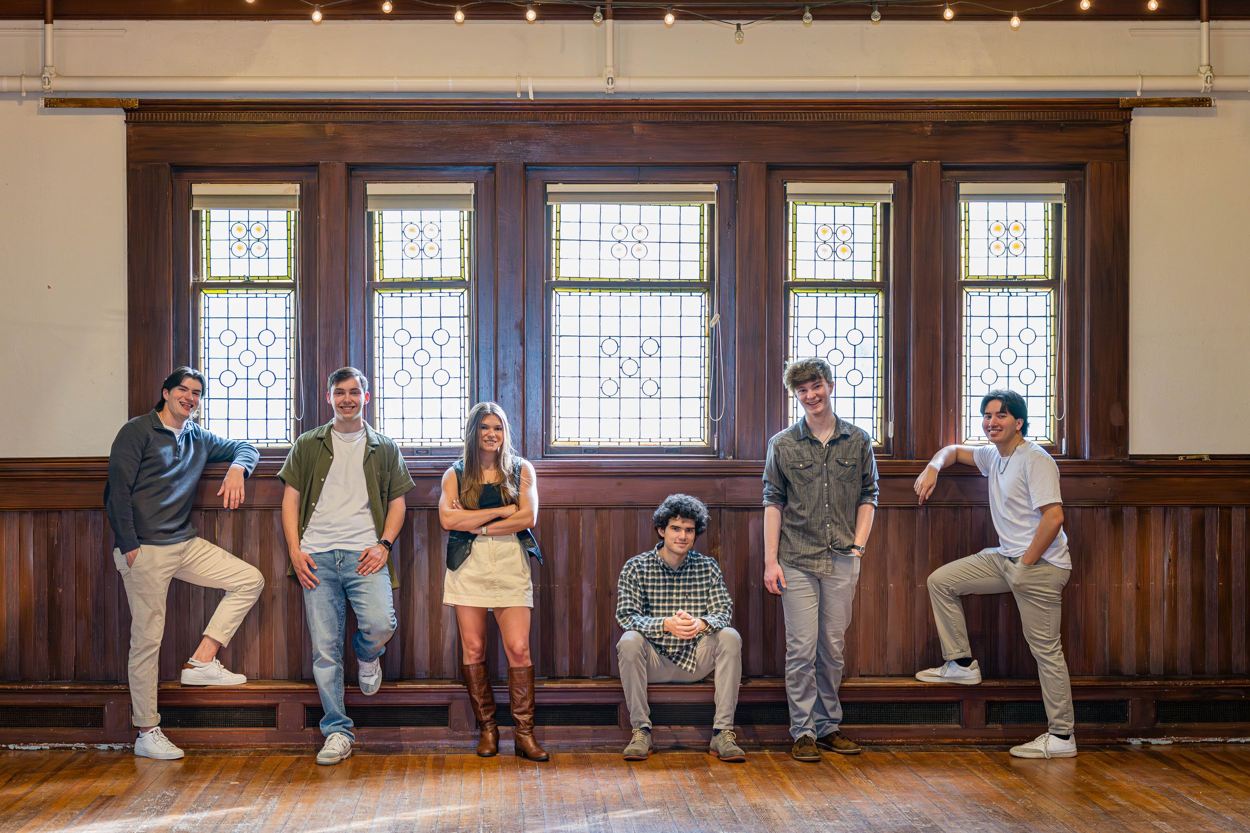 A panoramic sequence showing five people sitting against a wooden panel wall in various casual poses.