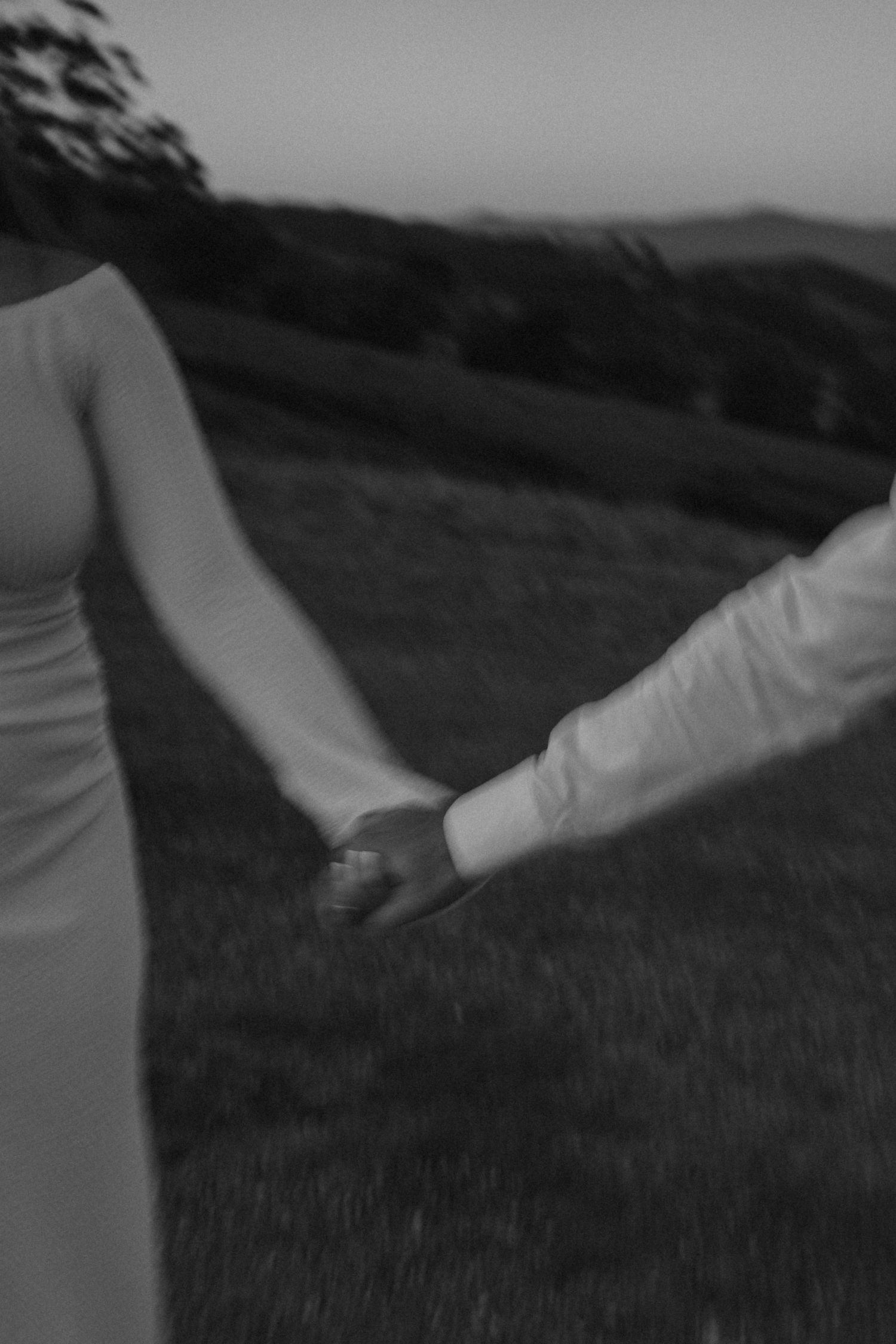 Black and white photo of a couple holding hands in a grassy field at dusk, showing only their arms and formal attire at Mt Lofty, Wonga Park