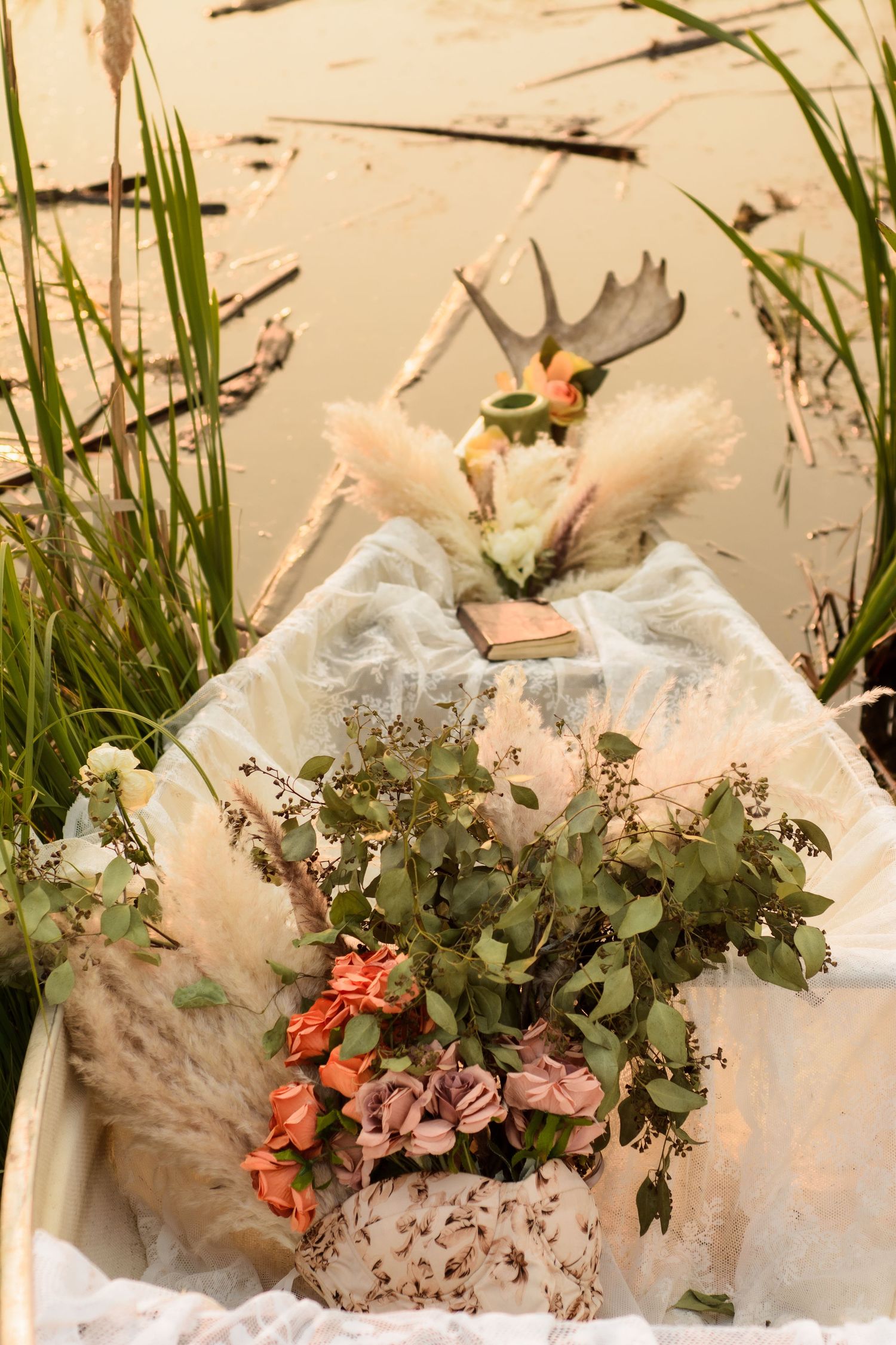 A romantic vintage-style wedding bouquet with dried flowers and greenery sits on white fabric near reeds and water.