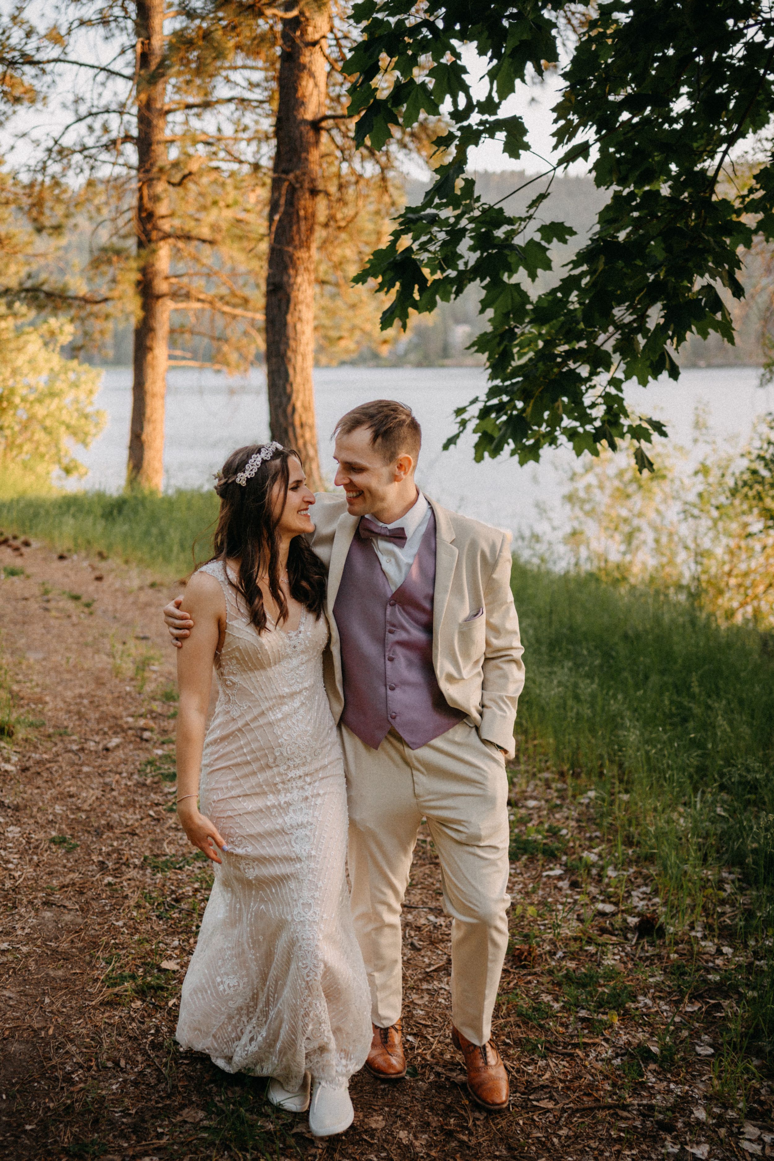 A couple in wedding attire walks together on a scenic woodland path at sunset by a lake.