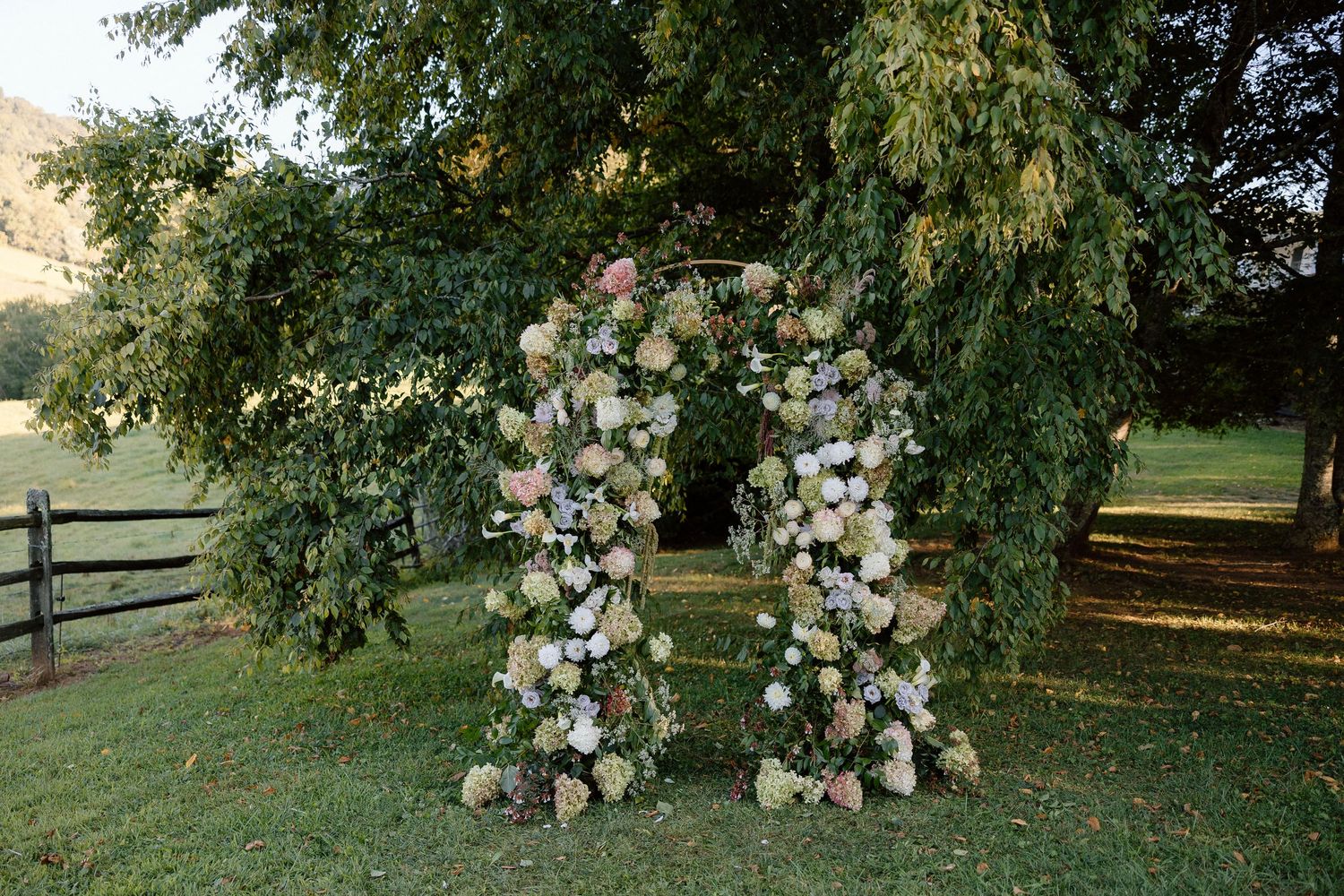 A rustic floral wedding arch decorated with white and blush roses stands on a grassy lawn with wooden fencing in the background.