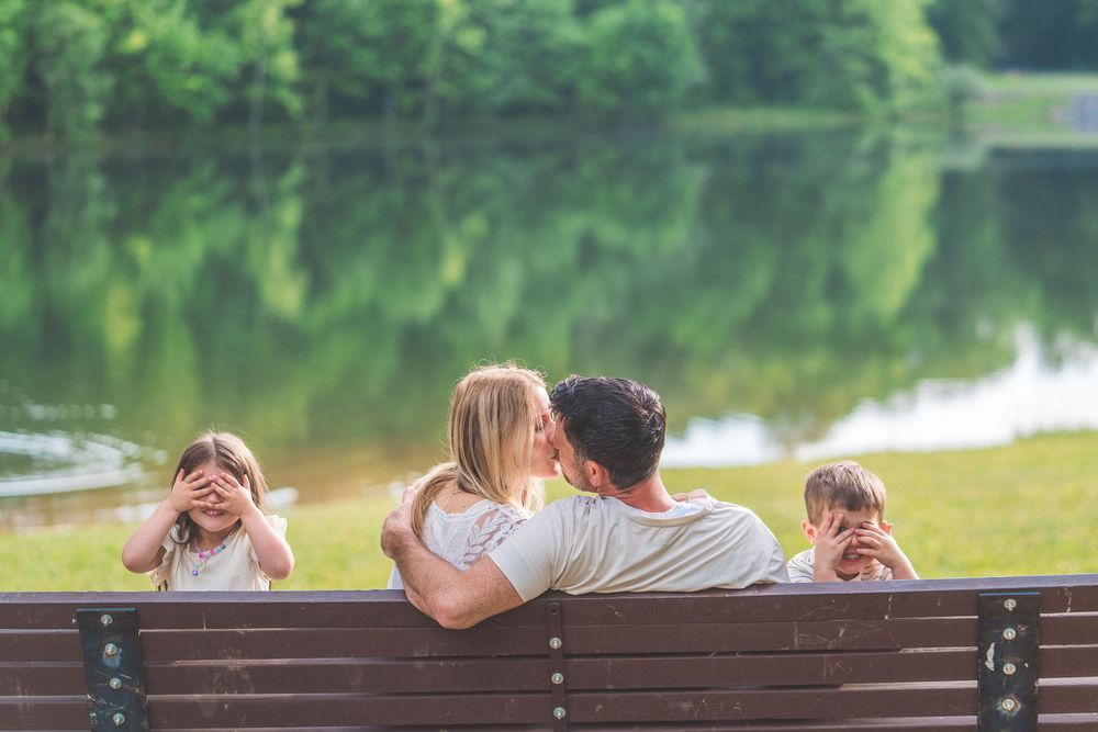 Family sitting together on park bench by serene lake with trees reflecting in water.