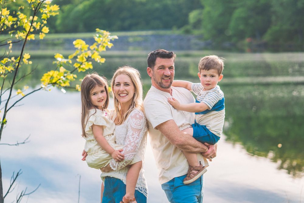 Family group portrait by lakeside with lush greenery and water reflections in background.