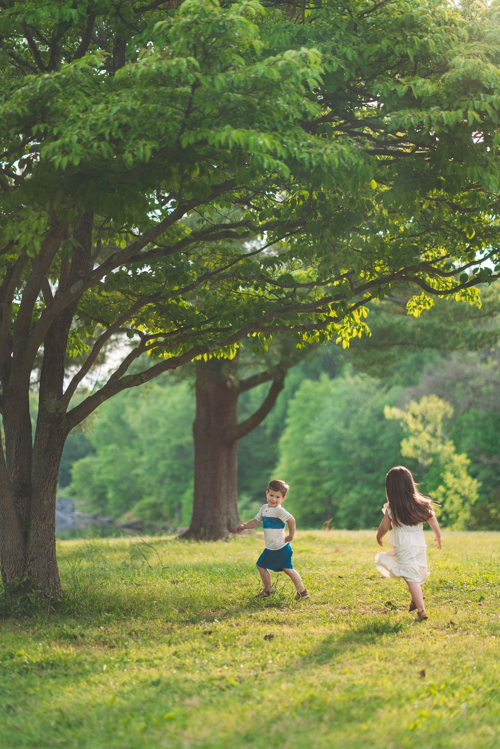 Two people running and playing under large shade trees in sunlit park.