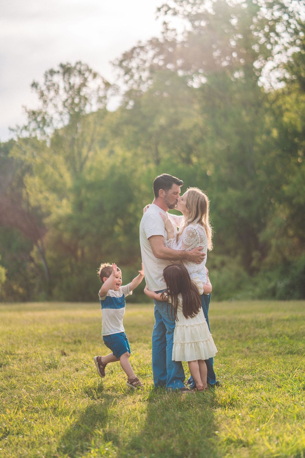 Family sharing intimate moment in sunlit field while children play around them.