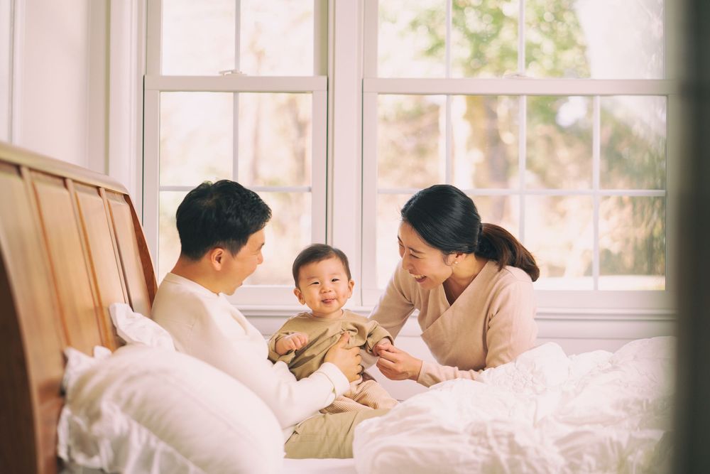 A family shares a tender moment together on a white bed by a sunlit window.