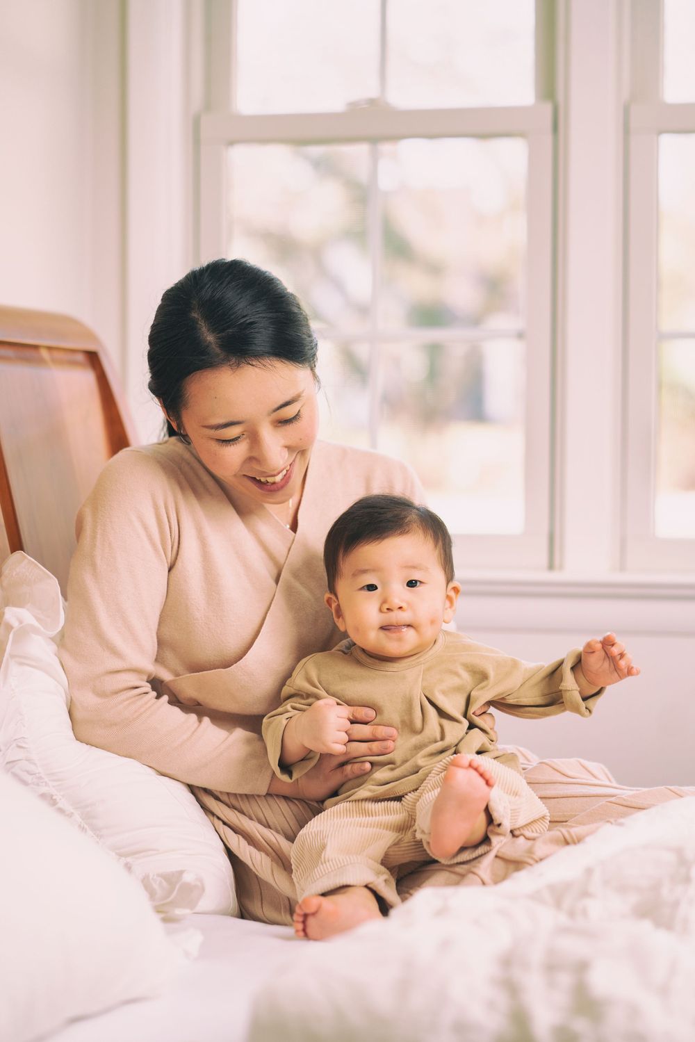 A parent and baby in matching beige outfits share a joyful moment on a white bed.
