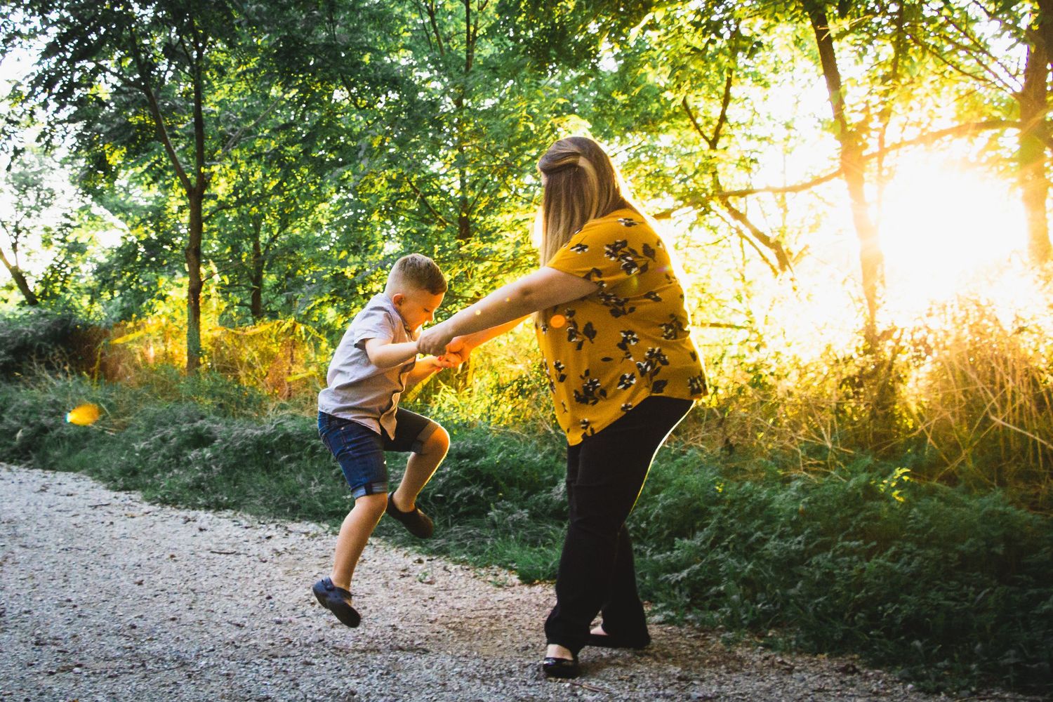 A mom and son playfully dancing together on a sunlit forest path with golden light streaming through the trees.