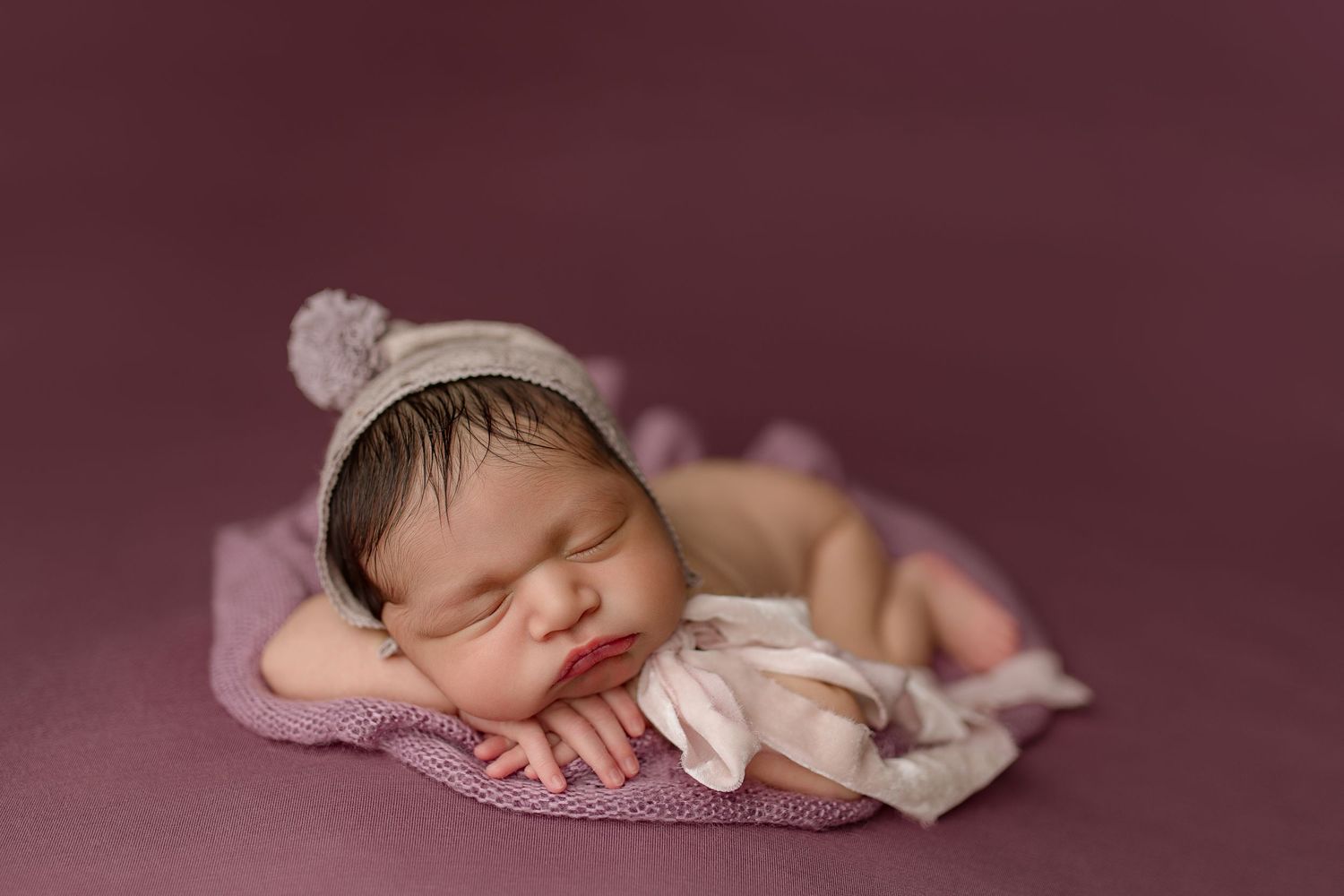 Sleeping newborn in white knit hat rests peacefully on pink background during photography session.
