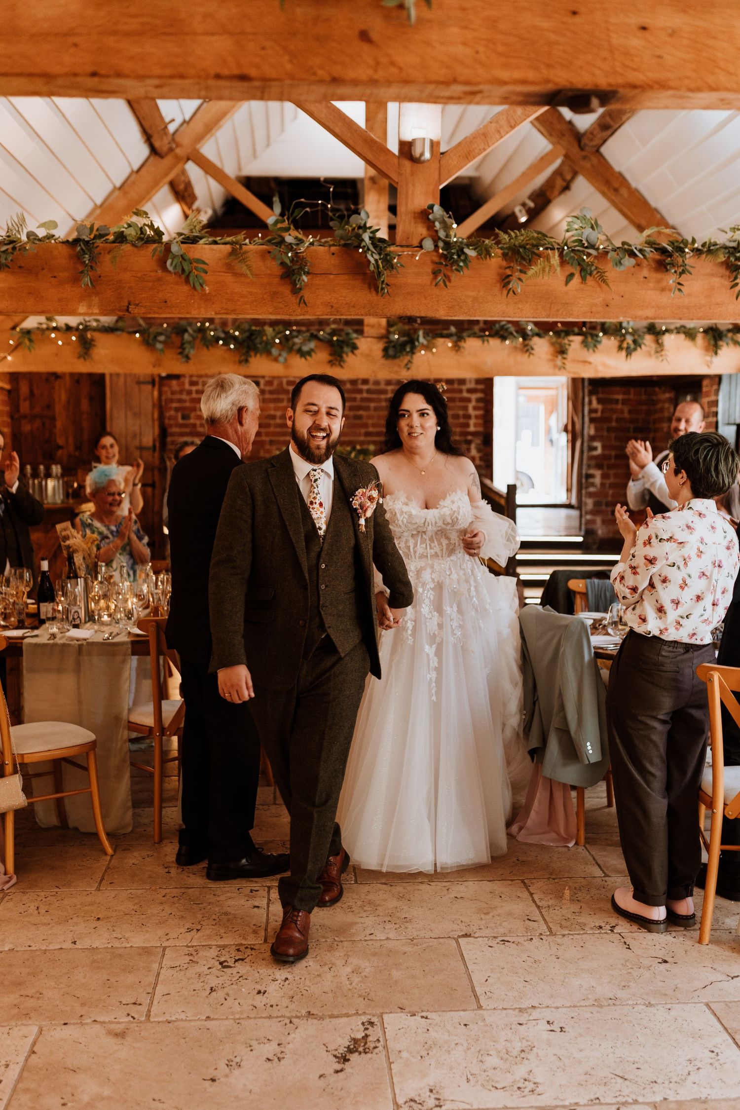 A rustic barn wedding reception venue with exposed wooden beams decorated with greenery garlands and elegant table settings.