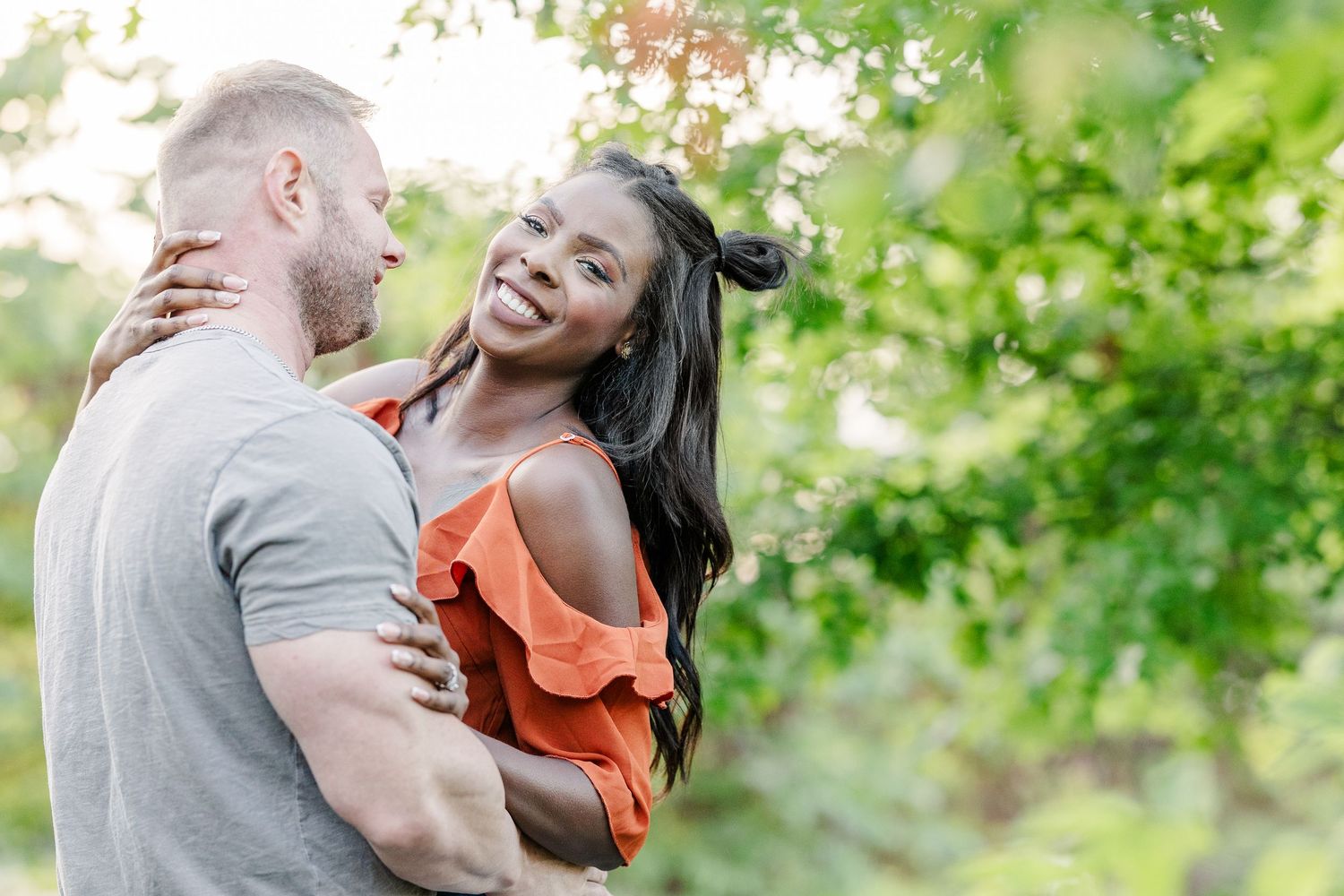 Couple in orange and gray outfits share intimate moments during an outdoor photoshoot with green foliage backdrop.