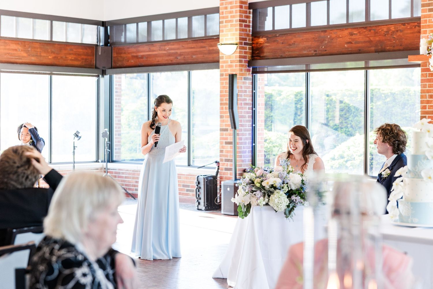 Someone gives a speech at an elegant indoor wedding reception with exposed brick walls and large windows.