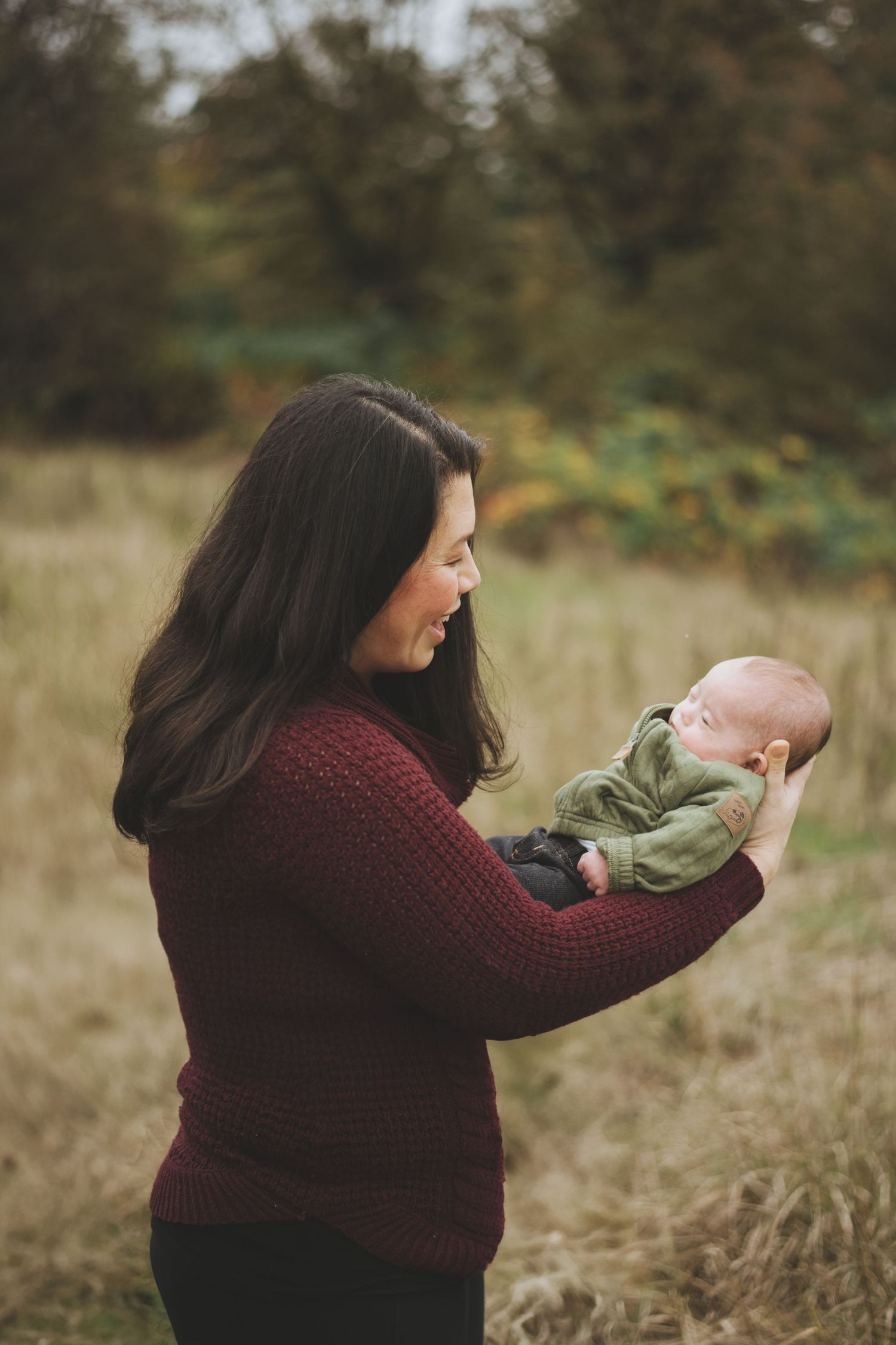 A woman in a burgundy sweater holds a baby wearing green in a sunlit autumn field.