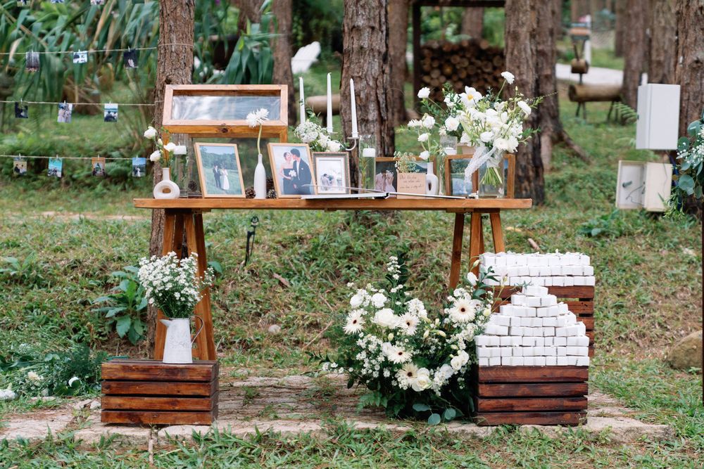 Rustic wooden display shelf with framed photos and white floral arrangements in an outdoor forest setting.