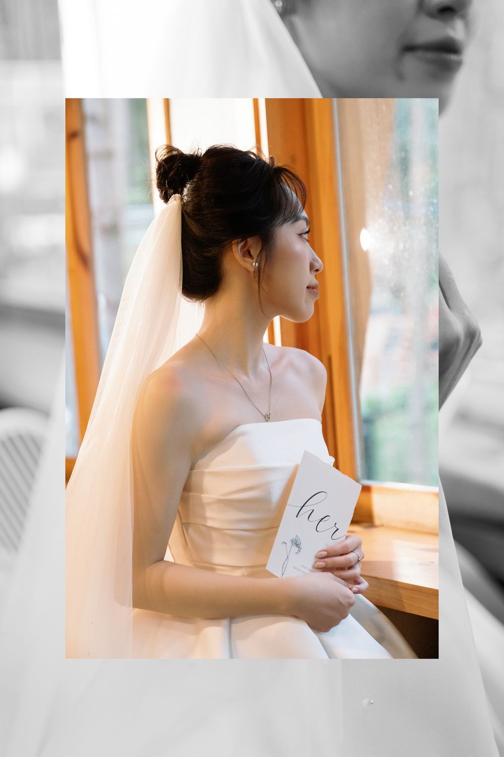 Elegant bride in strapless wedding dress holding invitation by window in soft lighting.