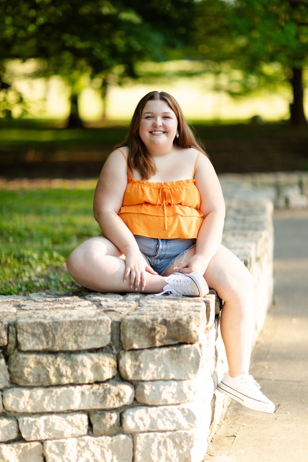 Person in orange top and denim shorts sitting casually on stone wall outdoors.