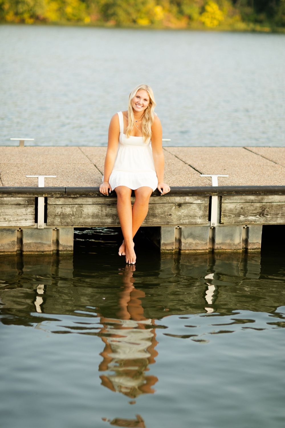 Person in white dress sits on wooden dock with feet dangling over calm lake water.