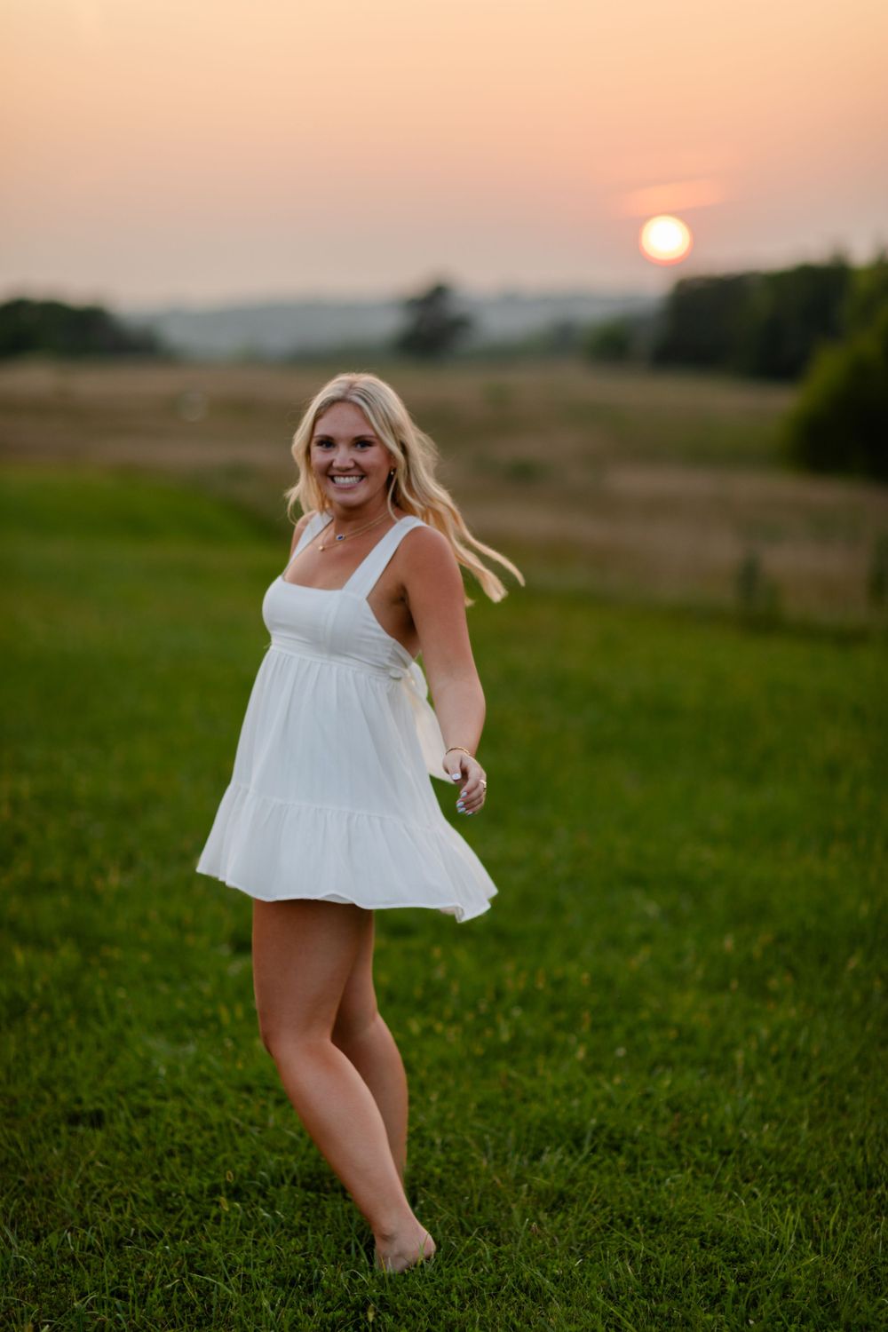 A senior girl in a white sundress twirls in a grassy field at sunset with warm orange sky in the background.