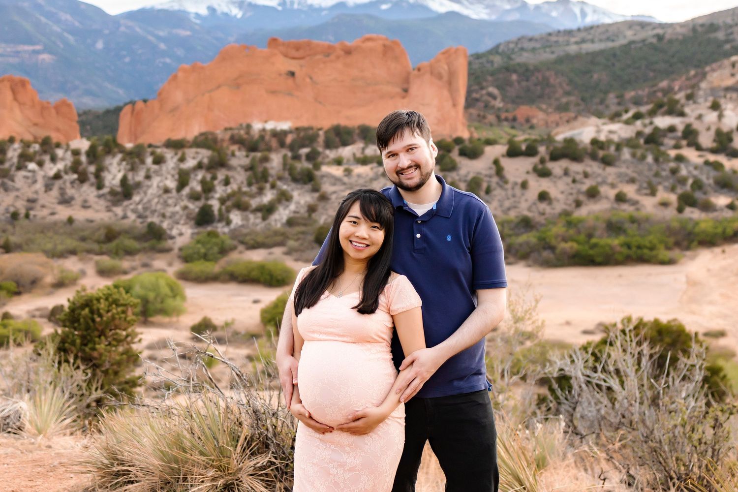Man stood behind pregnant woman, hugging her from behind, red mountains behind, she is wearing pink dress, holding bump