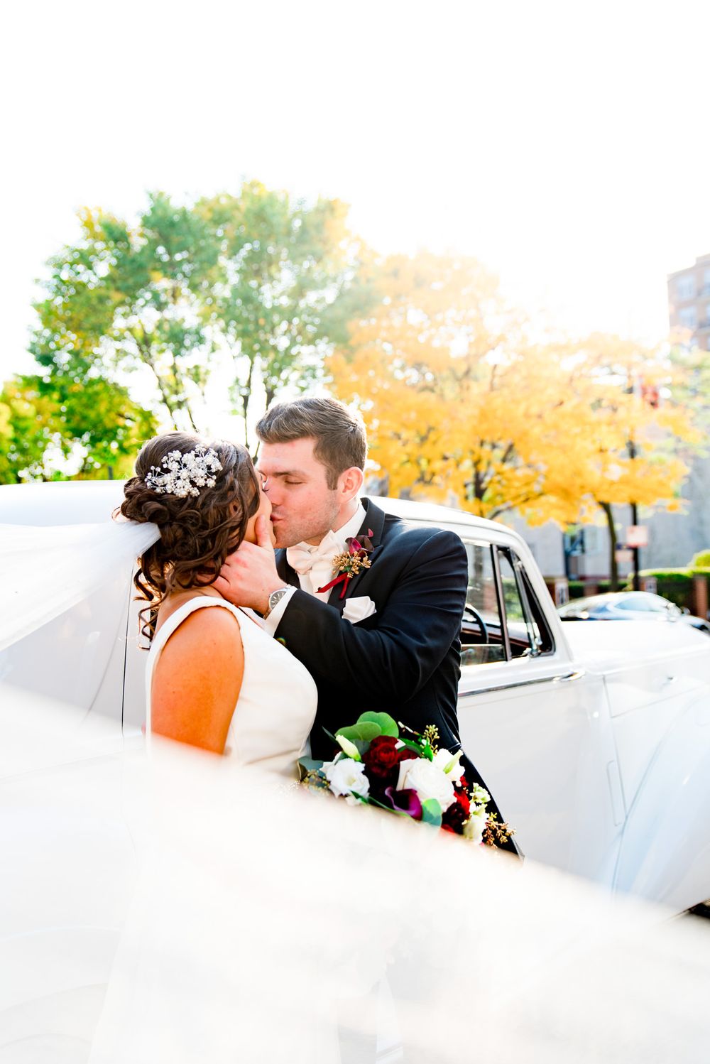colorful portrait of bride and groom kissing in front of antique white car