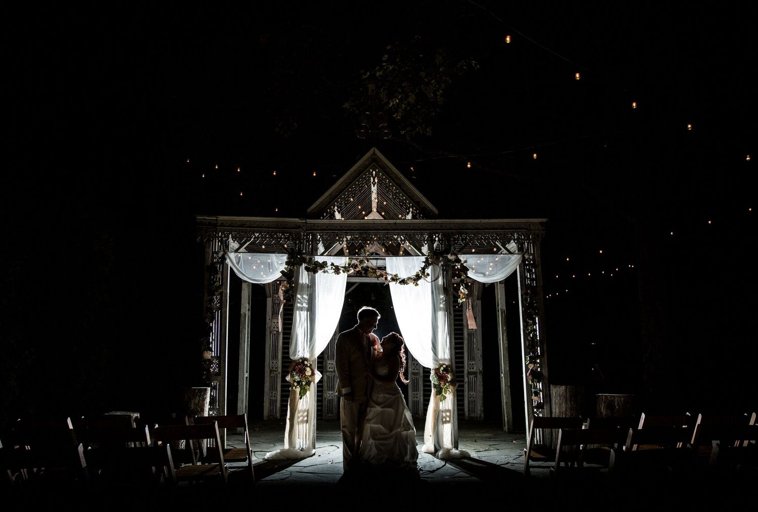Wedding ceremony backdrop decorated with white drapes and lights at night in a gazebo setting.