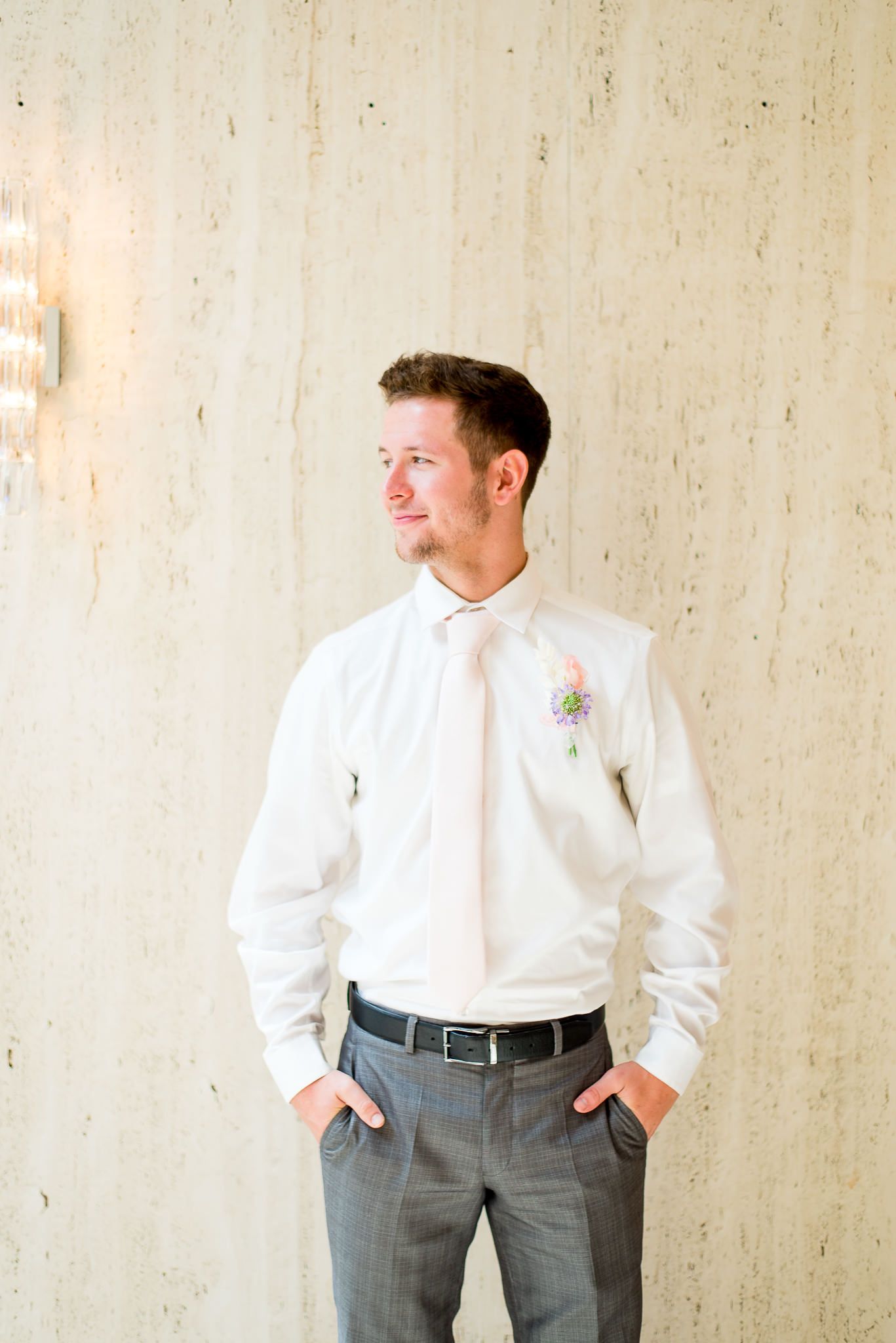 groom with blue eyes, light pink tie, and pink and purple boutonniere in front of marble wall at The Center Cincinnati