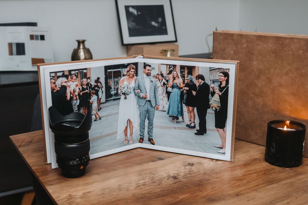 Wedding album open to show celebration photos with black candle and camera lens on wooden table.