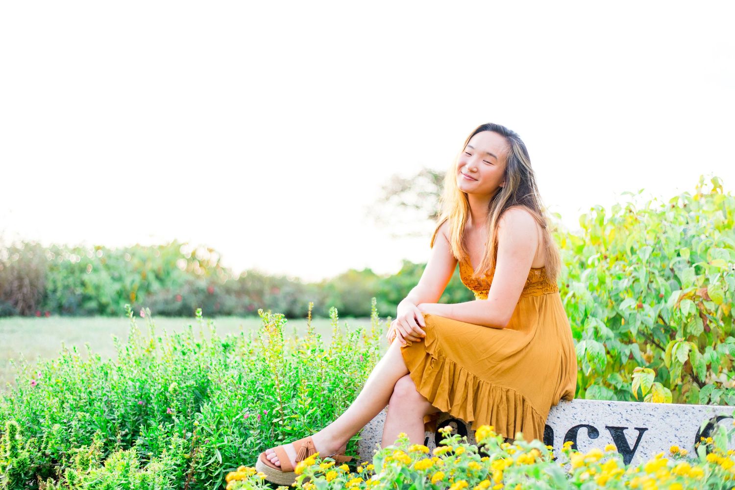 young woman sitting on a stone with legs crossed and smiling in a field of yellow flowers