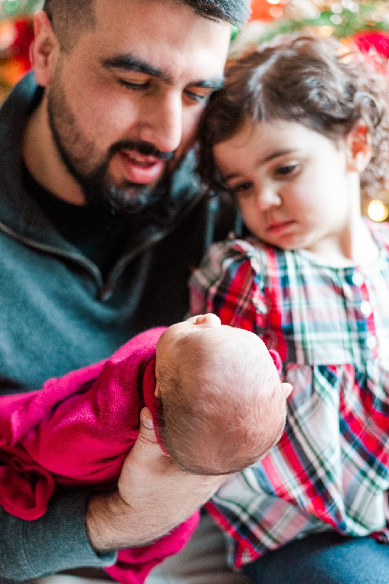 Lifestyle father and big sister holding newborn baby in pink wrap in front of Christmas tree in Pittsburgh, Pennsylvania