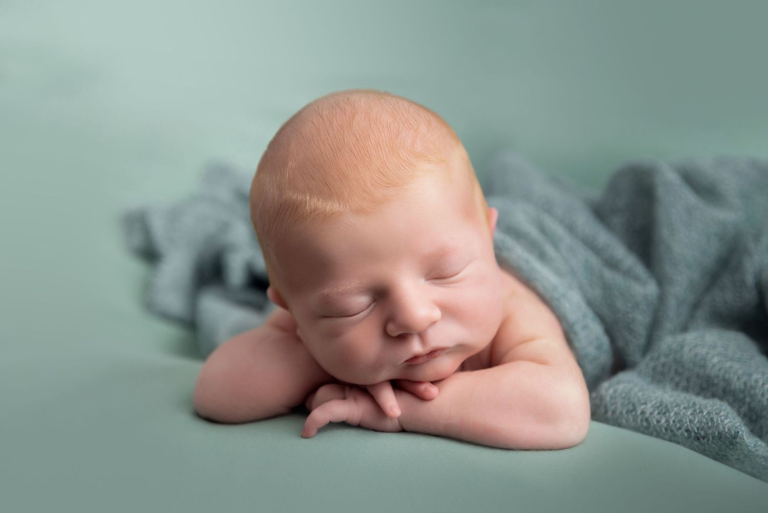 A sleeping newborn baby rests peacefully on their crossed arms with a soft gray blanket against a mint green background.