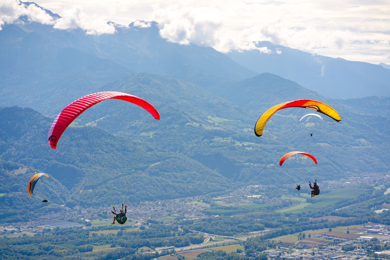 Multiple colorful paragliders flying over mountain valley with red and yellow canopies visible.