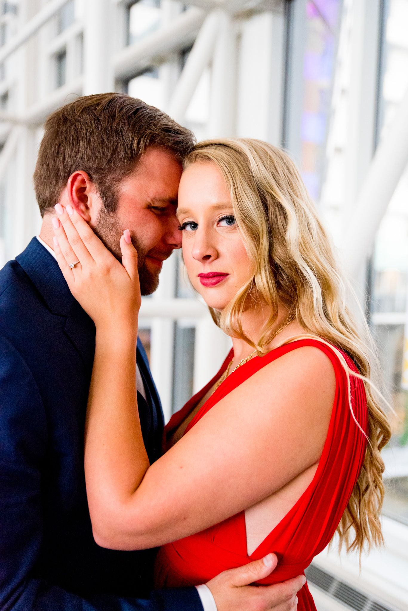 blonde woman in red dress looking at camera while fiance in navy suit rests on her forehead