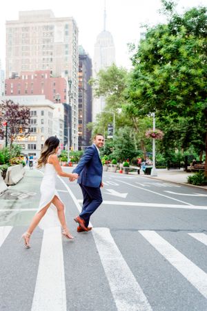 woman in white dress and man in suite holding hands running through street, view of Empire State Building behind them