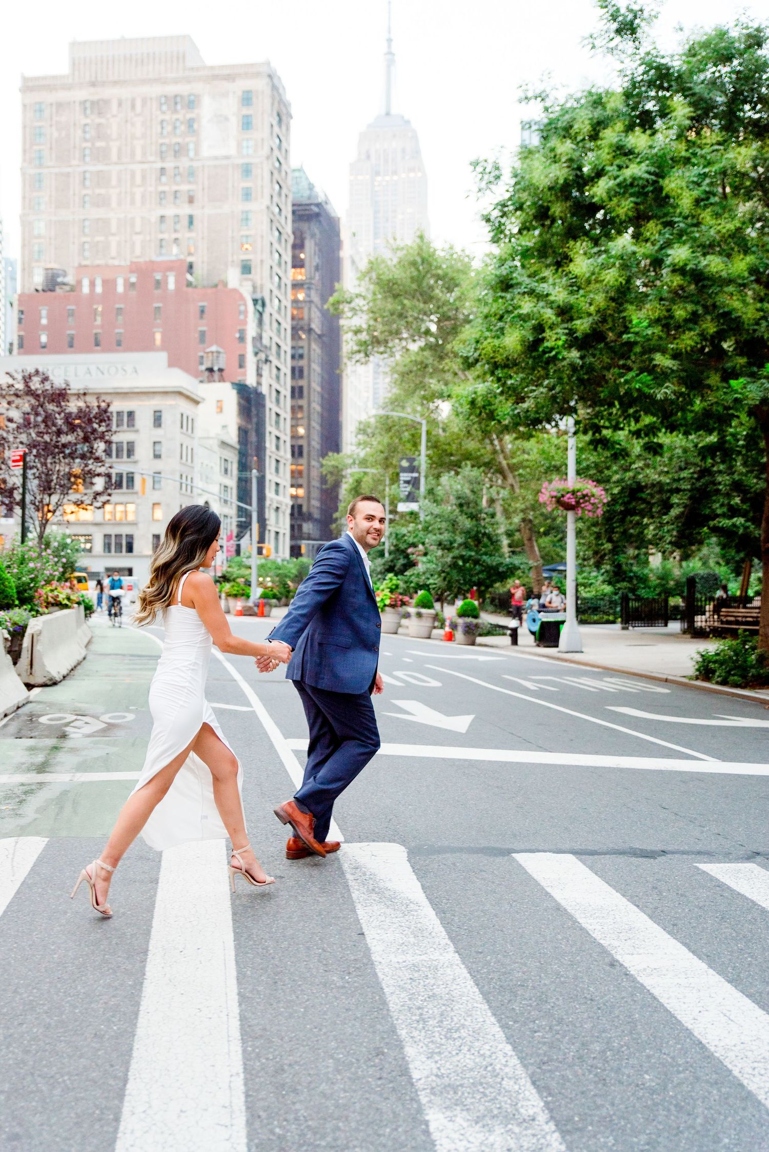 woman in white dress and man in suite holding hands running through street, view of Empire State Building behind them