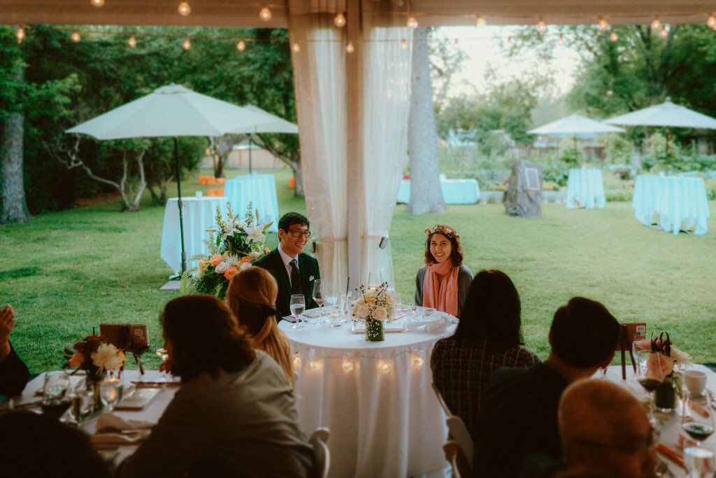 Outdoor wedding reception under white umbrellas with guests seated at round tables.