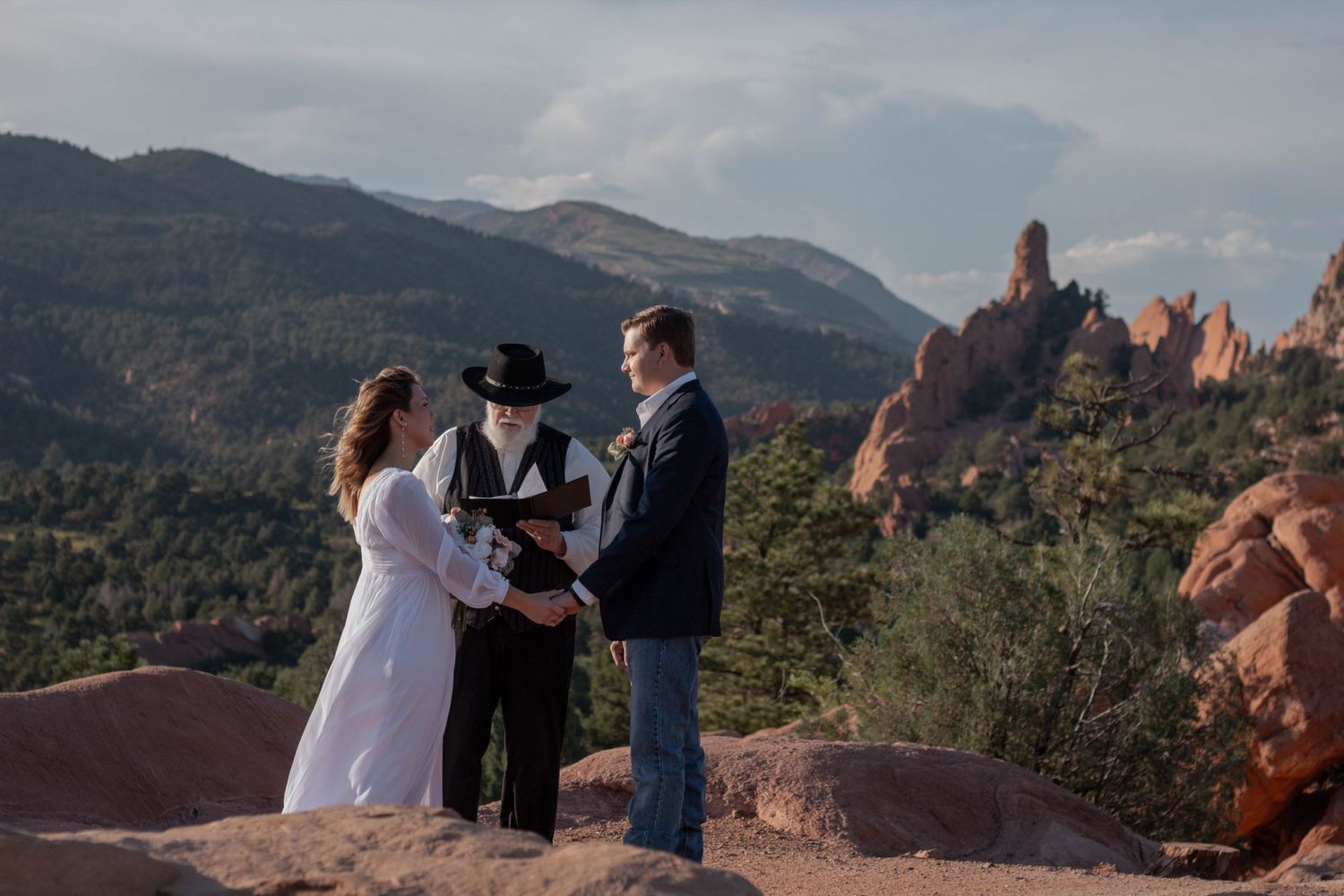 Wedding ceremony takes place on a scenic overlook with red rock formations and mountains in the background at Garden of the Gods. Elopement