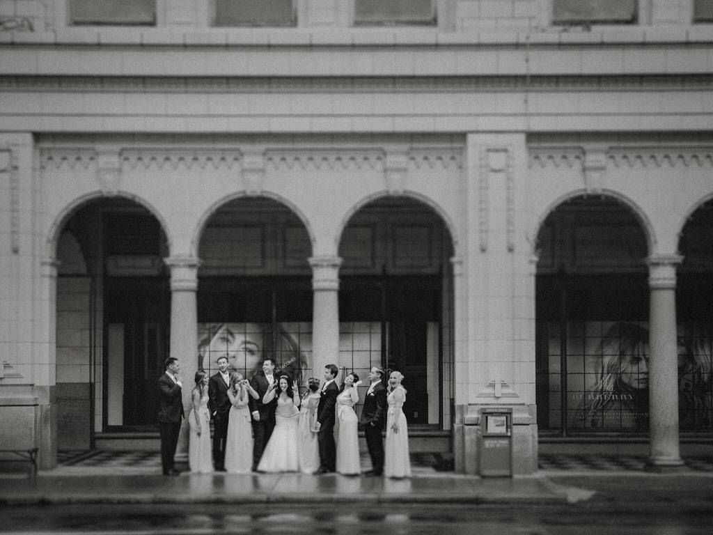 Classic black and white wedding party photo in front of an elegant building with arched columns.