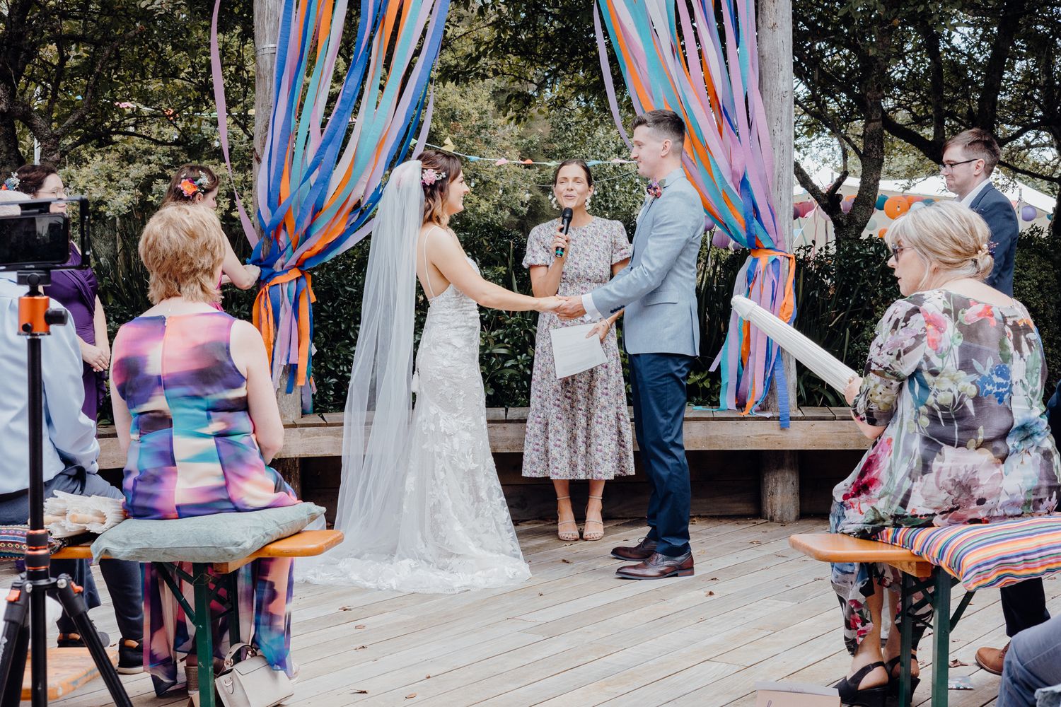 Outdoor wedding ceremony on wooden deck with colorful streamers and seated guests.