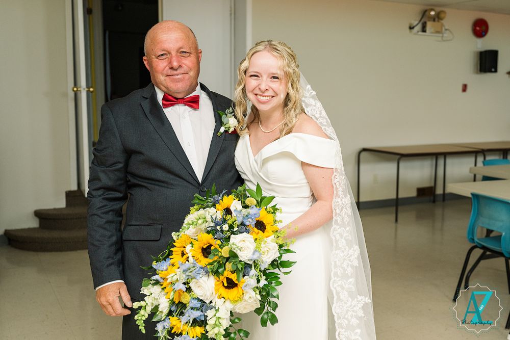 A wedding couple posing together indoors with a vibrant sunflower and white floral bouquet.