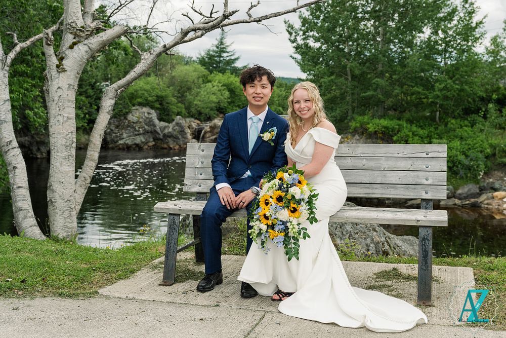 Newlyweds sit on bench by water with sunflower bouquet in scenic outdoor setting.