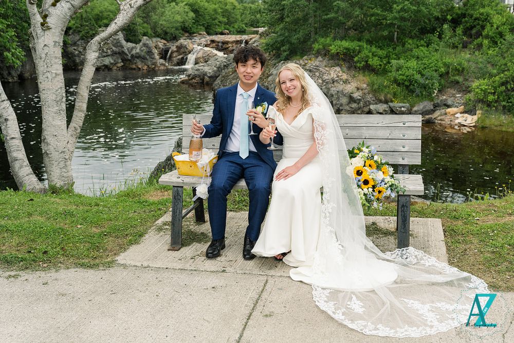 Wedding couple toasts with drinks while seated on bench near scenic waterfront.