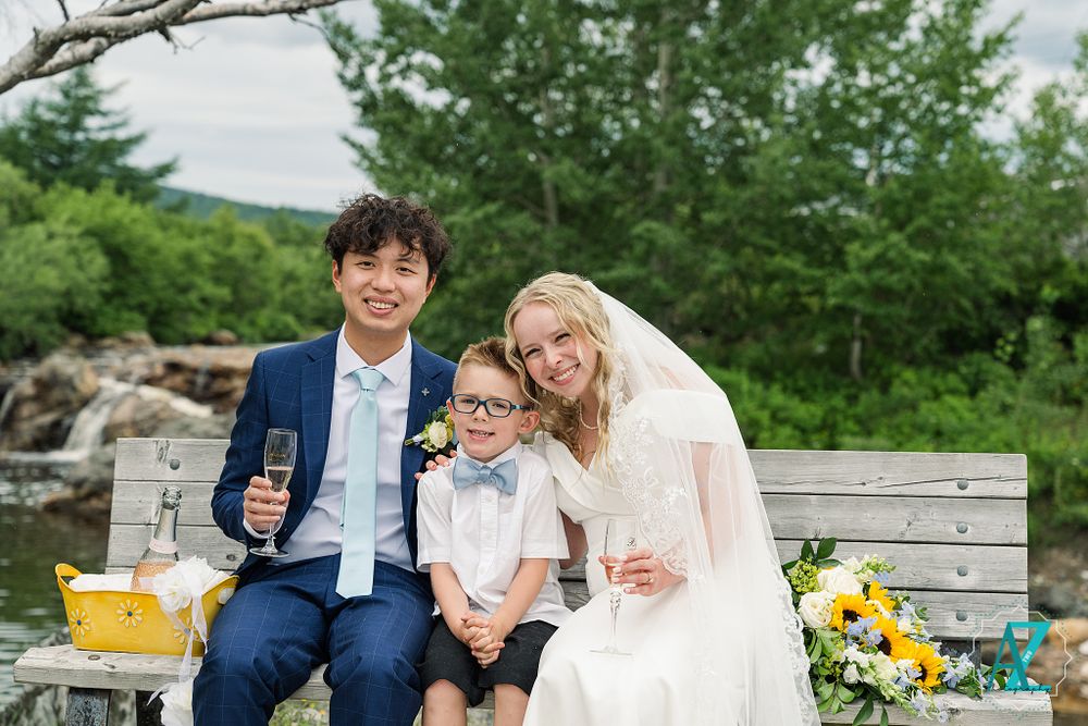 Wedding party enjoys drinks and celebration on wooden bench by waterfront.