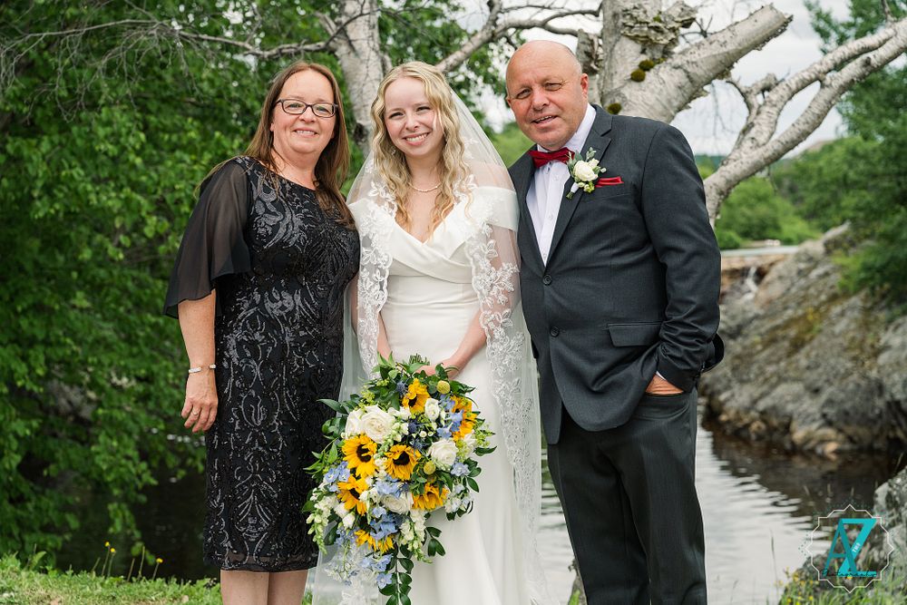 Wedding party poses with bouquet of sunflowers near rocky waterfront landscape.