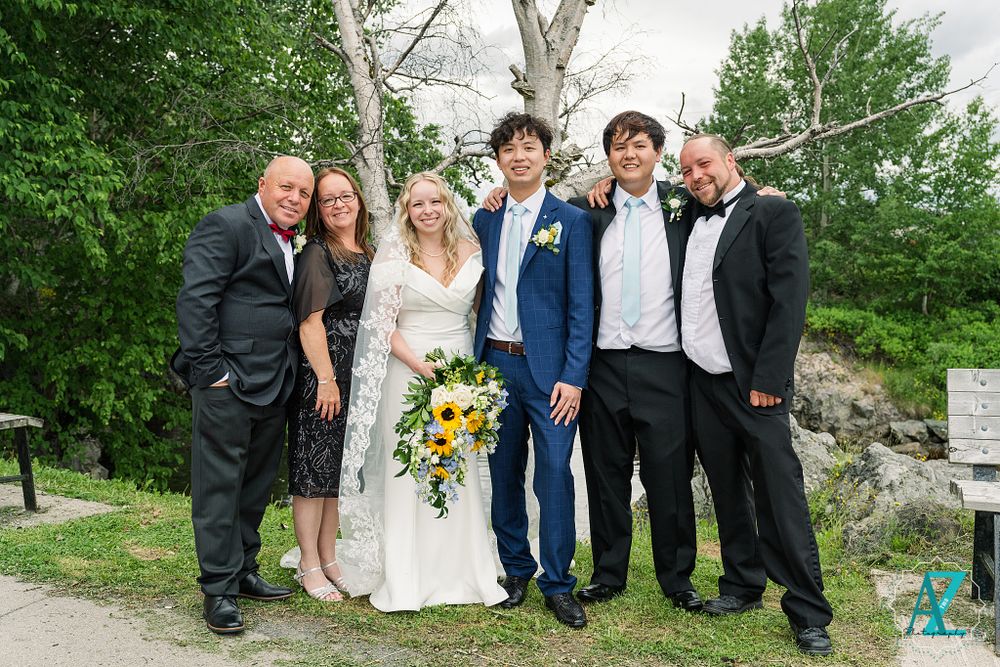 Wedding party poses together outdoors with bride holding vibrant sunflower bouquet against green trees and white birch.