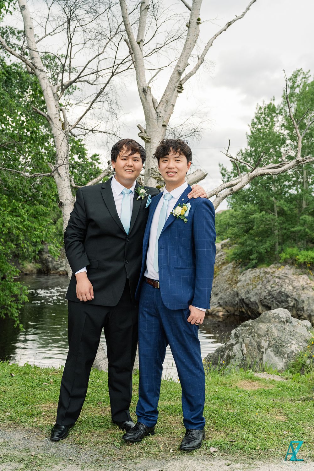 Two people in formal attire pose together by water with white birch trees in background.